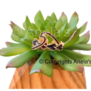 May include: Gold-tone ring with black enamel detailing in the shape of a wave. The ring is sitting on a green succulent plant.