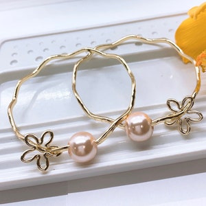 May include: Two gold-toned bangle bracelets with a wavy design, each with a pink pearl and a delicate flower charm.