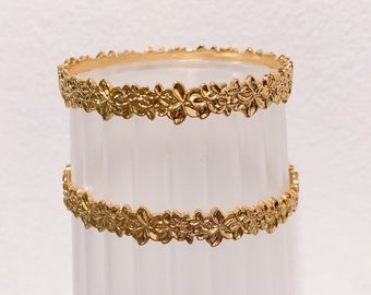 Puede incluir: Dos pulseras doradas con un diseño floral. Las pulseras están apiladas una encima de la otra.