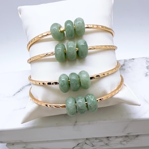 May include: Four gold-colored bangles, each adorned with three light green jade beads. The bangles have a hammered texture and are displayed on a white pillow. The background is a soft white.