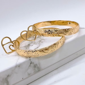 May include: Two gold-tone bangle bracelets with a floral design. The bracelets are stacked on top of each other and have a heart-shaped charm attached to one of the bracelets.