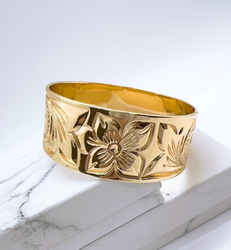 K&ouml;nnte beinhalten: Ein breiter goldfarbener Ring mit einem floralen Design. Der Ring ist aus Metall und hat eine glatte, polierte Oberfl&auml;che.