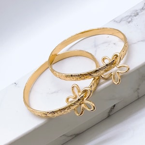 May include: Two gold bangles with intricate floral designs and a flower-shaped clasp. The bangles are stacked on top of each other.