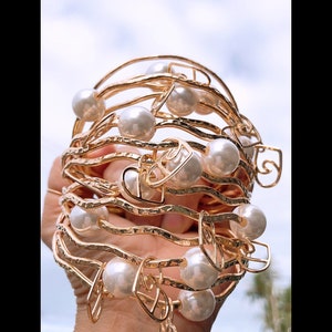 May include: A set of gold-toned bangle bracelets with a textured finish and pearl accents. The bracelets are stacked on top of each other and feature a heart-shaped design.