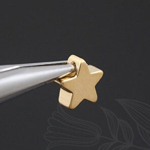 May include: A gold star charm with a small loop at the top. The charm is being held by a pair of tweezers.