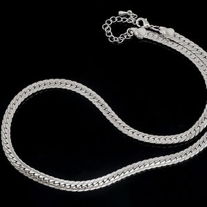 Puede incluir: Un collar de cadena de plata con un diseño delicado y tejido. La cadena está hecha de pequeños eslabones entrelazados que crean un aspecto suave y fluido.
