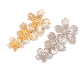 May include: Two hair clips, one in a gold tone and one in a silver tone, each featuring a cluster of three five-petalled flowers with textured, grooved surfaces and detailed centres, arranged in a linear, ascending size pattern.