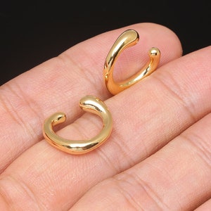 Könnte beinhalten: Zwei goldfarbene Ohrringe, die jeweils die Form eines leicht unregelmäßigen, gekrümmten Kreises haben. Die Ohrringe werden auf einer Hand gezeigt, mit einem Ohrring an jedem Finger.