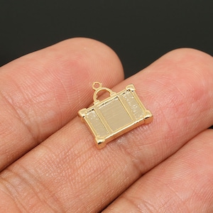 May include: Gold-coloured metal charm shaped like a vintage suitcase with a handle. The charm is small and detailed.