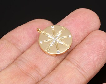 May include: A gold-coloured pendant featuring a compass design. The compass rose is embellished with small, clear crystals. The pendant has a loop at the top for a chain. The background is black.
