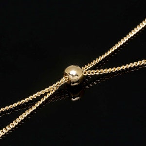 Op de afbeelding: Een gouden ketting met een ronde kraal in het midden. De ketting bestaat uit twee strengen die met de kraal zijn verbonden.