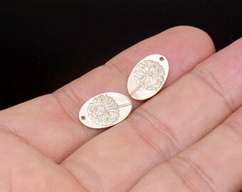 May include: Two gold-colored oval pendants with a dandelion design. Each pendant has a small hole at the top for stringing. The pendants are held in a person's hand against a black background. These pendants are suitable for jewelry making.