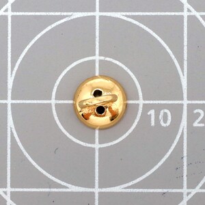 May include: A gold button with two holes and a loop on the back. The button is round and has a shiny finish. The button is on a grey surface with white grid lines and the number 10.