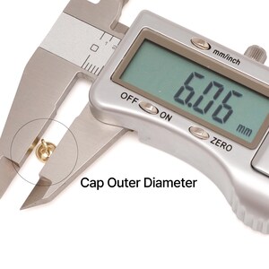 May include: A digital caliper measuring a small gold-coloured cap. The digital display shows 6.06 mm. The text "Cap Outer Diameter" is printed below the cap. The caliper is silver and the background is white.
