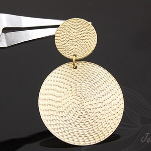 Könnte beinhalten: Goldfarbene Ohrringe mit einem strukturierten, kreisförmigen Design. Die Ohrringe bestehen aus zwei Kreisen, einer kleiner als der andere, die durch eine kleine Schlaufe verbunden sind.