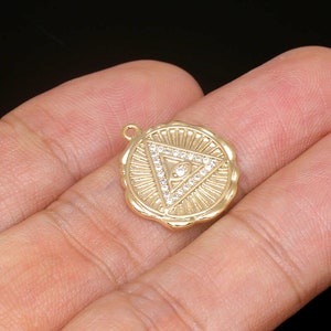 Puede incluir: Pendiente de encanto dorado con un diseño de triángulo que presenta un solo ojo en el centro, rodeado de diamantes de imitación transparentes. El pendiente tiene un borde texturizado, como de rayos de sol.