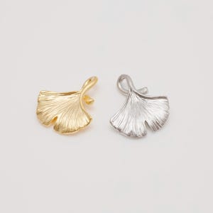 May include: Two metal ginkgo leaf pendants, one gold-toned and one silver-toned, are displayed against a white background. The pendants have a textured surface and a loop at the top for hanging. The leaves are approximately 1 inch wide.