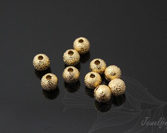 May include: A group of 10 gold-colored round beads with a textured surface. The beads are scattered on a black surface.