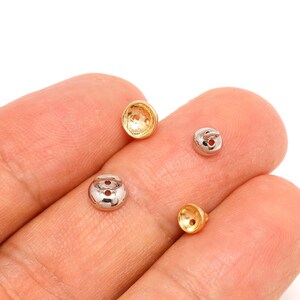 May include: Four small, metallic earring backs in gold and silver tones. The earring backs are round with a small hole in the centre and a slightly flared base. The backs are displayed against a light background.