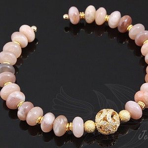 May include: A gold bracelet with a sunstone bead design and a decorative gold charm. The bracelet is made with round, flat beads in shades of pink and brown.