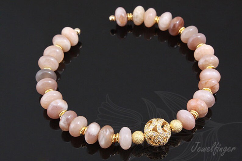 May include: A gold bracelet with a sunstone bead design and a decorative gold charm. The bracelet is made with round, flat beads in shades of pink and brown.
