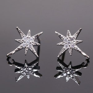 Pode incluir: Um par de brincos de estrela prateados com pedras de zircônia cúbica transparentes. Os brincos têm um design pontudo e são montados em um poste prateado.