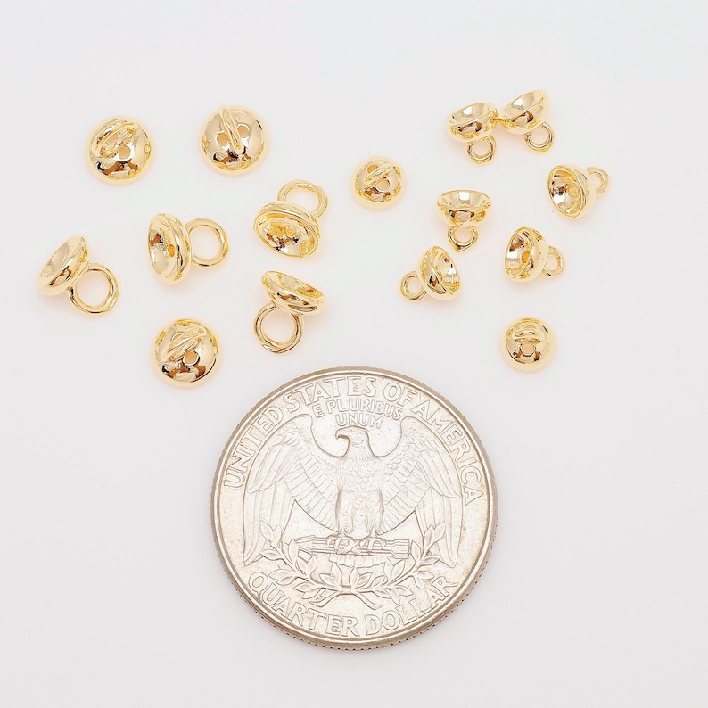 May include: A selection of small, gold-coloured jewellery findings, including bell-shaped beads and components with loops. A U.S. quarter coin is included for size reference. The findings are scattered on a white surface.