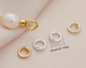 May include: Close-up of jewellery components, including a large white pearl, gold-coloured clasps, and small silver and gold-coloured rings. The rings are approximately 5mm or 0.19in in diameter. The background is a light pink fabric.