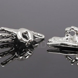 Puede incluir: Dos colgantes de plata con forma de mano haciendo el signo de OK. Los colgantes son detallados y tienen un acabado liso y pulido.