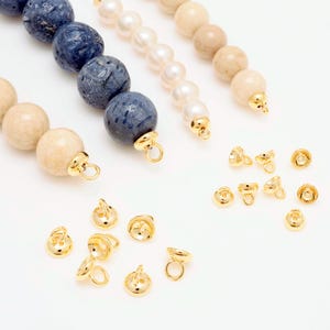 May include: An overhead view of jewellery components. Strands of beads in blue, white, and beige are visible, alongside several gold-coloured bead caps. The beads are round and smooth, while the bead caps have a loop for attaching to other jewellery pieces.