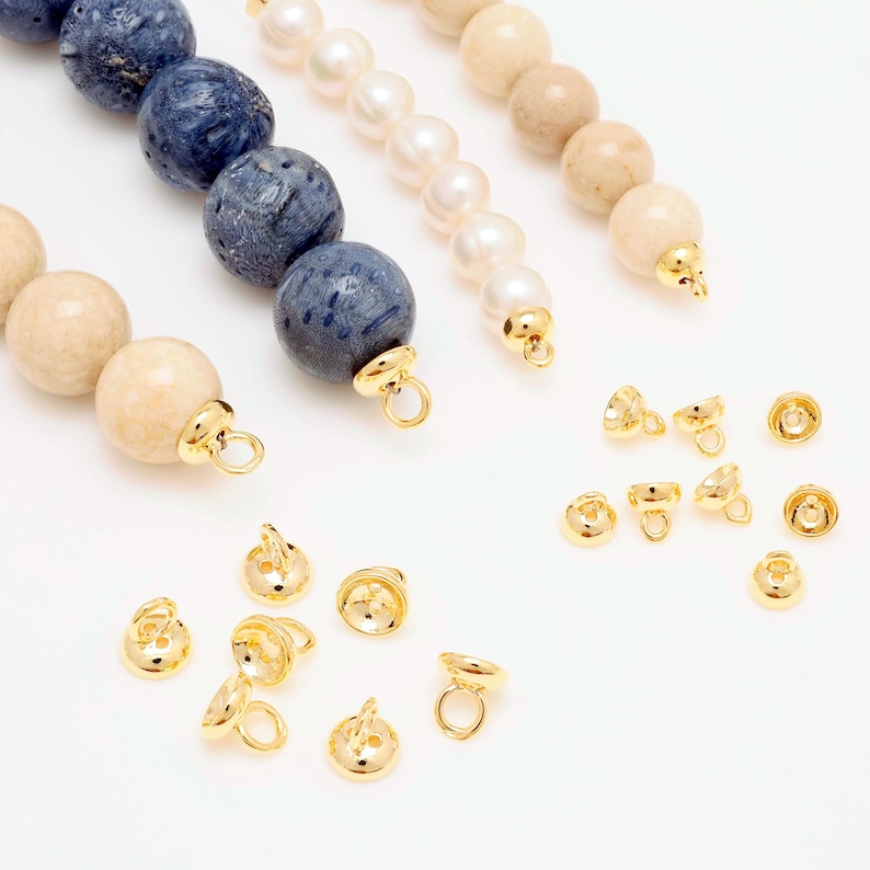May include: An overhead view of jewellery components. Strands of beads in blue, white, and beige are visible, alongside several gold-coloured bead caps. The beads are round and smooth, while the bead caps have a loop for attaching to other jewellery pieces.