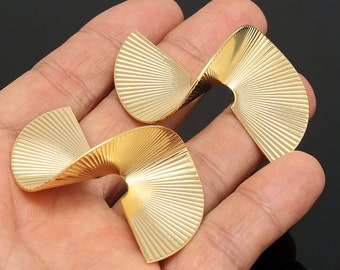May include: Two gold-coloured, abstract earrings with a fan-like design. Each earring has a curved, ridged surface that catches the light. The earrings are displayed against a dark background.