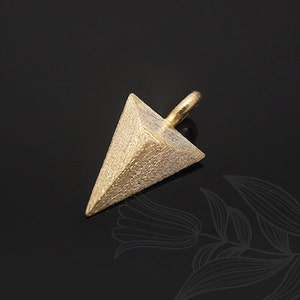 Puede incluir: Un dije de metal de color dorado en forma de pirámide con una superficie texturizada. El dije tiene un bucle en la parte superior para sujetarlo a un collar o pulsera.