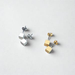 Könnte beinhalten: Zwei Paar quadratische Ohrstecker. Ein Paar ist silberfarben, das andere goldfarben. Die Ohrringe haben ein schlichtes, modernes Design mit einem kleinen Würfel, der an einem Stecker und einem Butterfly-Verschluss befestigt ist.