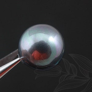 May include: A dark gray pearl with iridescent green and purple hues. The pearl is being held by a pair of tweezers.