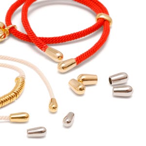 May include: A bright red cord and a cream-colored cord, both featuring polished gold-tone metal end caps. Several loose, bullet-shaped metal cord ends in gold and silver tones are scattered nearby, showcasing their smooth, rounded, and hollow design.