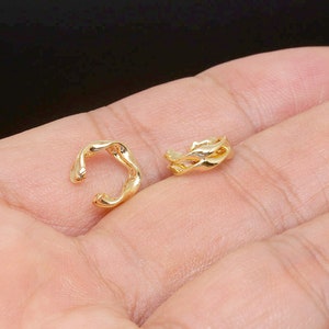 Könnte beinhalten: Zwei goldene Ohrclips, einer mit einem einfachen gedrehten Design und der andere mit einem komplizierteren, wellenförmigen Design. Die Ohrclips werden auf einer Hand gezeigt, bereit zum Tragen.