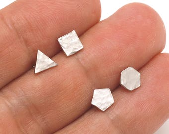 May include: A collection of four silver-colored stud earrings in geometric shapes: a triangle, a square, a hexagon, and a pentagon. The earrings have a textured, hammered finish. The earrings are small and delicate, suitable for everyday wear.