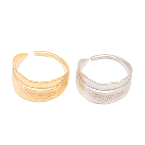 Puede incluir: Dos anillos ajustables con forma de pluma, uno dorado y otro plateado, se muestran sobre un fondo blanco. Los anillos presentan un diseño detallado de pluma y una banda abierta y ajustable. Los anillos probablemente son de metal.