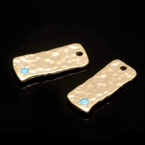 Op de afbeelding: Twee rechthoekige metalen labels in goudkleur met een gehamerde textuur en een klein turquoise blauwe edelsteen in het midden van elk label.