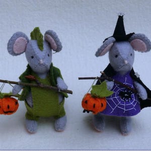 May include: Two handmade felt mice dressed for Halloween. One mouse wears a green outfit and carries an orange pumpkin. The other mouse is dressed as a witch in a purple dress with a spiderweb design and a black cape, also carrying a pumpkin.