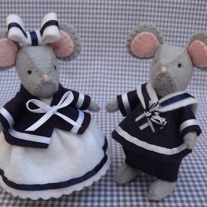 Peut inclure: Deux souris en feutre faites à la main, vêtues de tenues de marin. L'une porte une robe blanche avec une bordure et une veste bleu marine. L'autre porte un costume de marin bleu marine. Les deux ont des oreilles roses et des nœuds blancs.