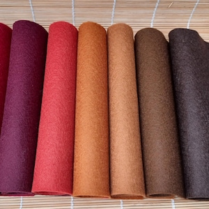 May include: A collection of rolled felt sheets in various colors, including burgundy, purple, red, orange, brown, and dark brown. The felt has a textured surface and is arranged on a bamboo mat.