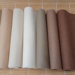 Puede incluir: Una colección de hojas de fieltro enrolladas en varios colores neutros, incluyendo beige, crema y marrón. El fieltro tiene una apariencia texturizada, y los rollos están dispuestos uno al lado del otro, mostrando la gama de tonos disponibles.