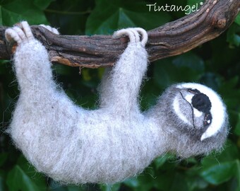 May include: A fuzzy, light grey needle-felted sloth figurine hanging from a brown tree branch. The sloth has a white face mask, a black nose, and black eye patches. The text Tintangel is visible in the upper right corner of the image.