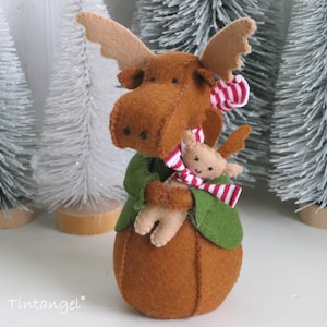 Op de afbeelding: Een handgemaakt vilten elandfiguur met een bruin lichaam, geweien en een groene jas. De eland houdt een kleinere eland vast met een rood-witte gestreepte sjaal. De achtergrond bevat kleine witte kerstbomen.
