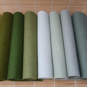 May include: A selection of rolled felt sheets in shades of green, white, and grey. The felt is displayed on a bamboo mat, highlighting the different colours and textures. The felt appears soft and pliable, suitable for crafts.