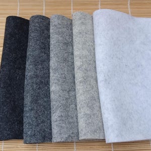 Puede incluir: Muestras de tela de fieltro en tonos grises y blanco. Las muestras están dispuestas en fila, mostrando los diferentes tonos, desde el carbón oscuro hasta el gris claro y el blanco. El fieltro tiene un aspecto suave y texturizado.