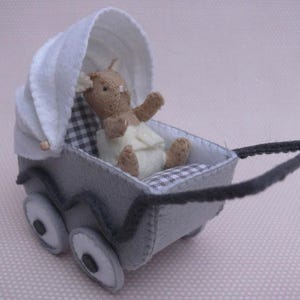 Op de afbeelding: Een miniatuur grijze vilten kinderwagen met witte accenten, een witte kap en grote wielen. Binnenin zit een kleine knuffelbeer met een luier op een geruite stof. De kinderwagen heeft een zwart handvat.