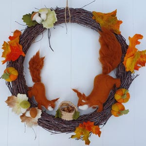 Può includere: Una ghirlanda autunnale fatta a mano con una base circolare in vite. È adornata con scoiattoli di feltro, zucche e foglie colorate nei toni dell'arancione, giallo e marrone. È incluso anche un piccolo cestino.